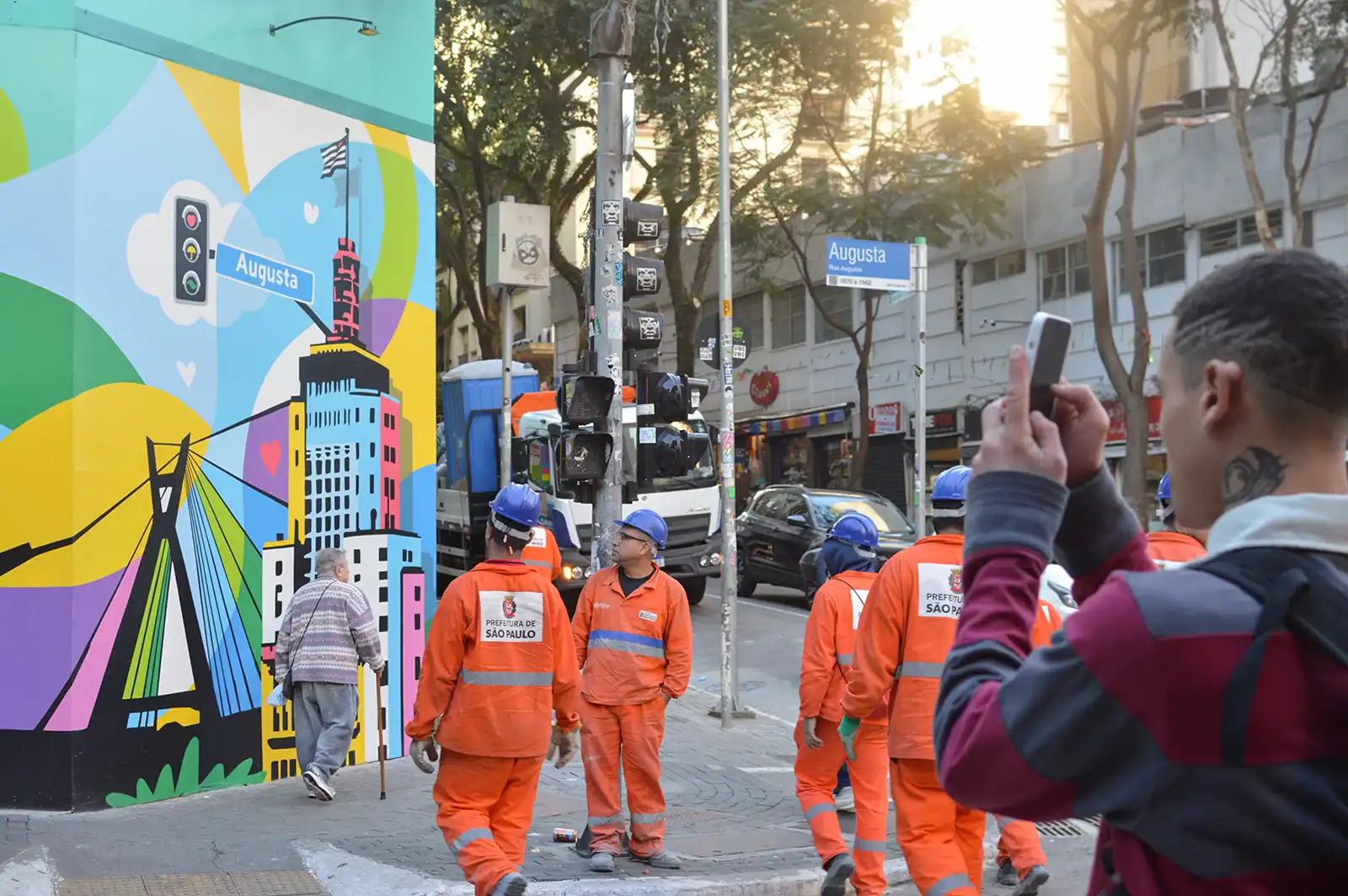 Trabalhadores e pedestres circulam pela esquina enquanto um visitante fotografa o mural