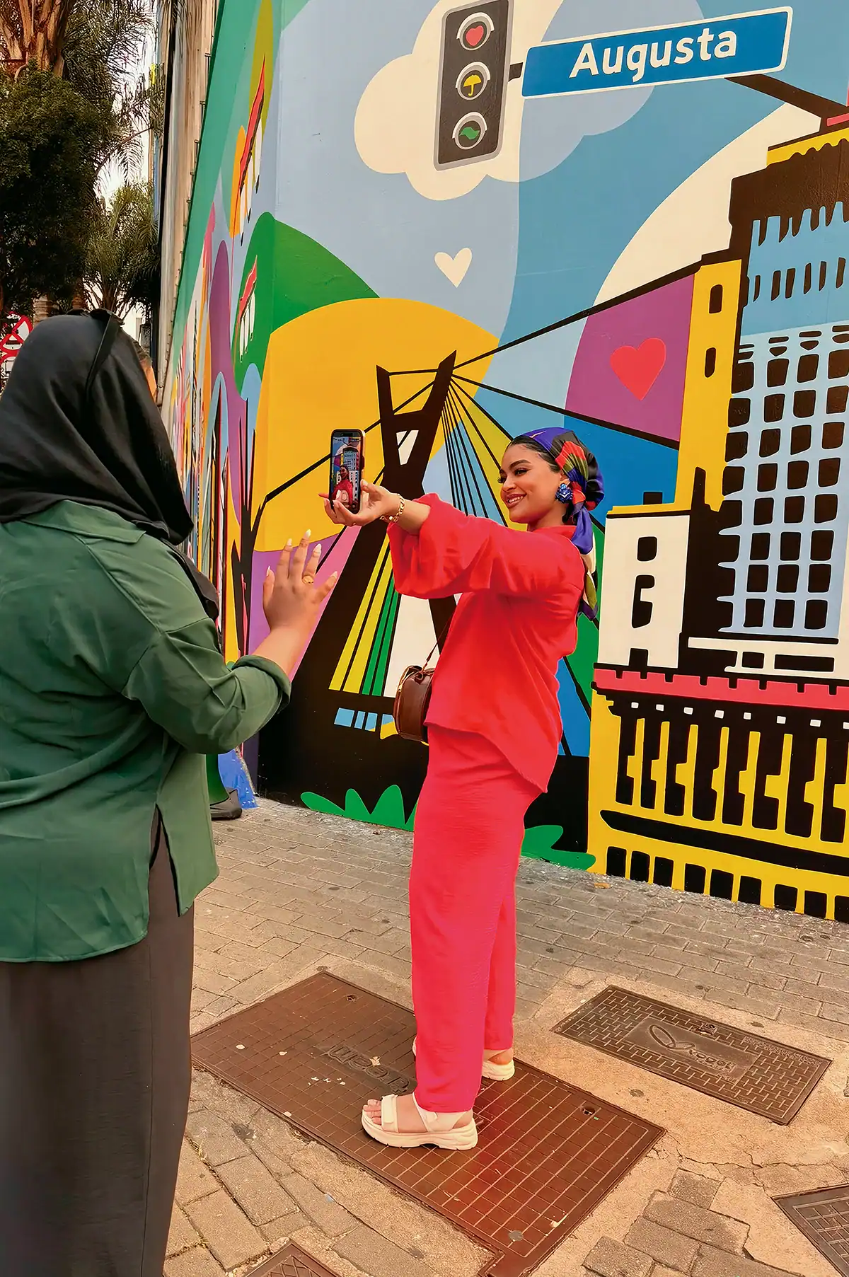 Mulher de vermelho faz uma selfie diante da obra colorida enquanto outra visitante acompanha a cena